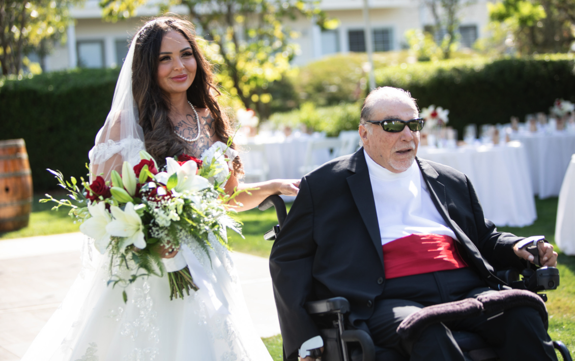 A bride in a while dress is escorted down the aisle by a man in a wheelchair